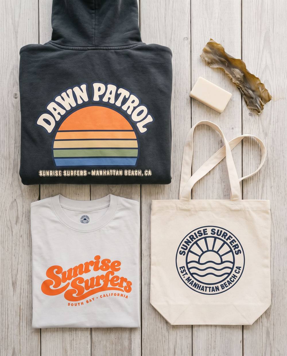 Sunrise Surfers Collection 01 flat lay: the Dawn Patrol hoodie, Script Tee and canvas tote on weathered wood.