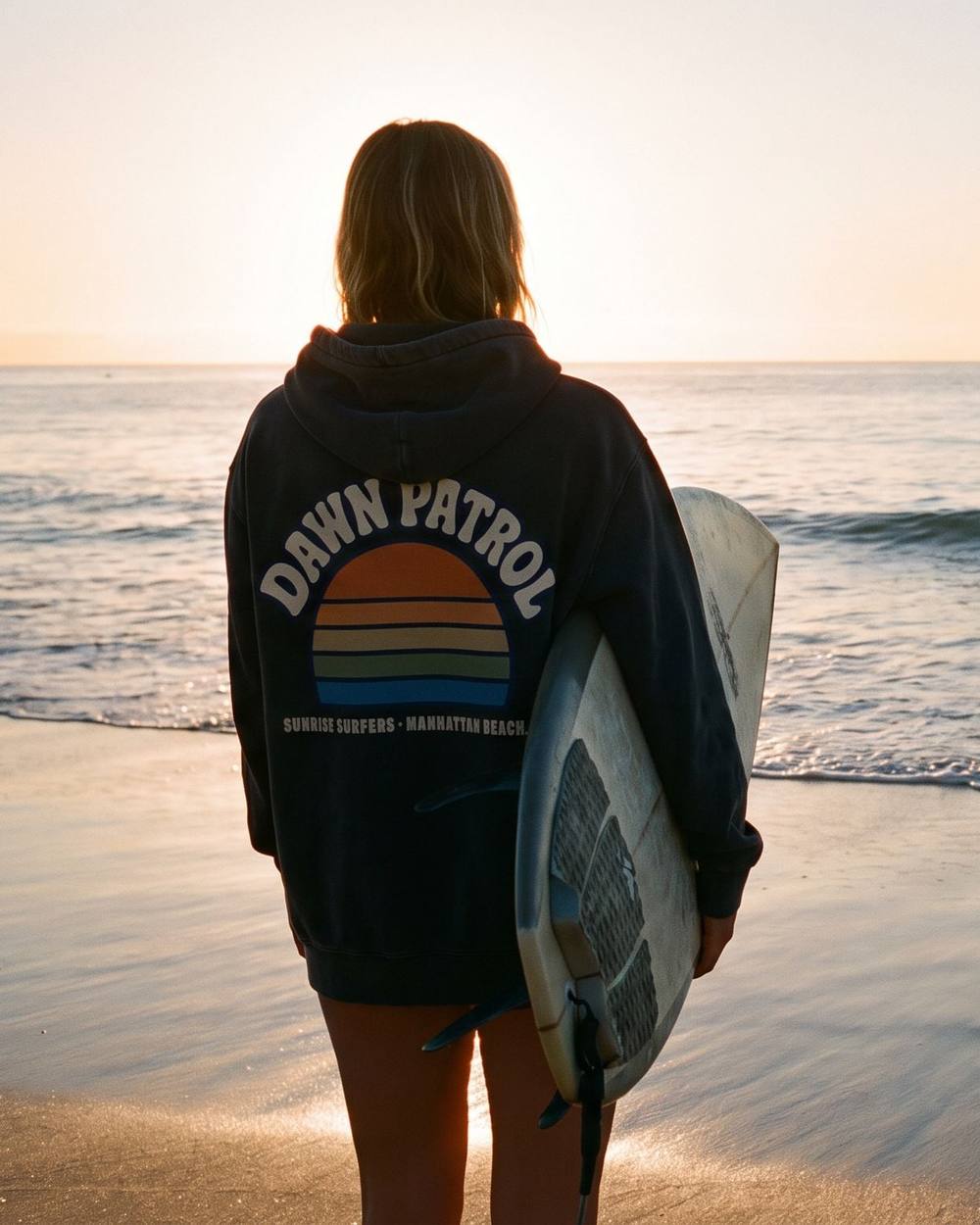Surfer on the sand at sunrise in the Sunrise Surfers Dawn Patrol hoodie, showing the striped sunrise graphic across the back.