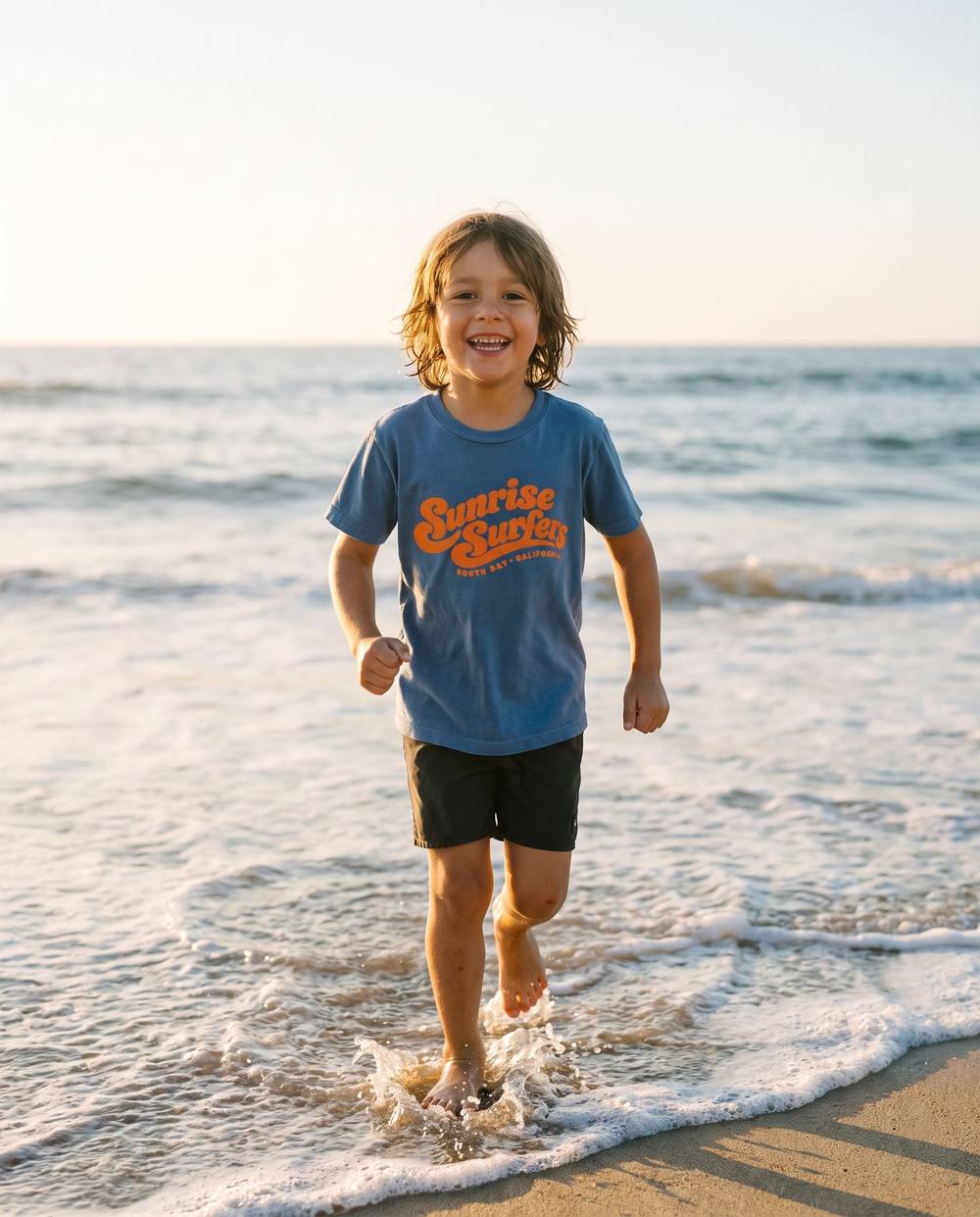 Child running through the shorebreak at sunrise in the Sunrise Surfers Kids Tee in Blue Jean.